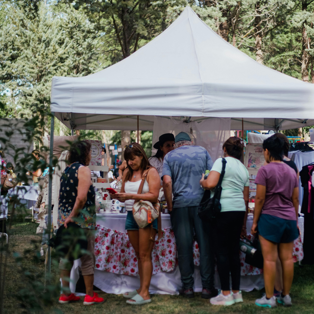 Shoppers browsing under a white vendor tent at an outdoor market surrounded by trees.